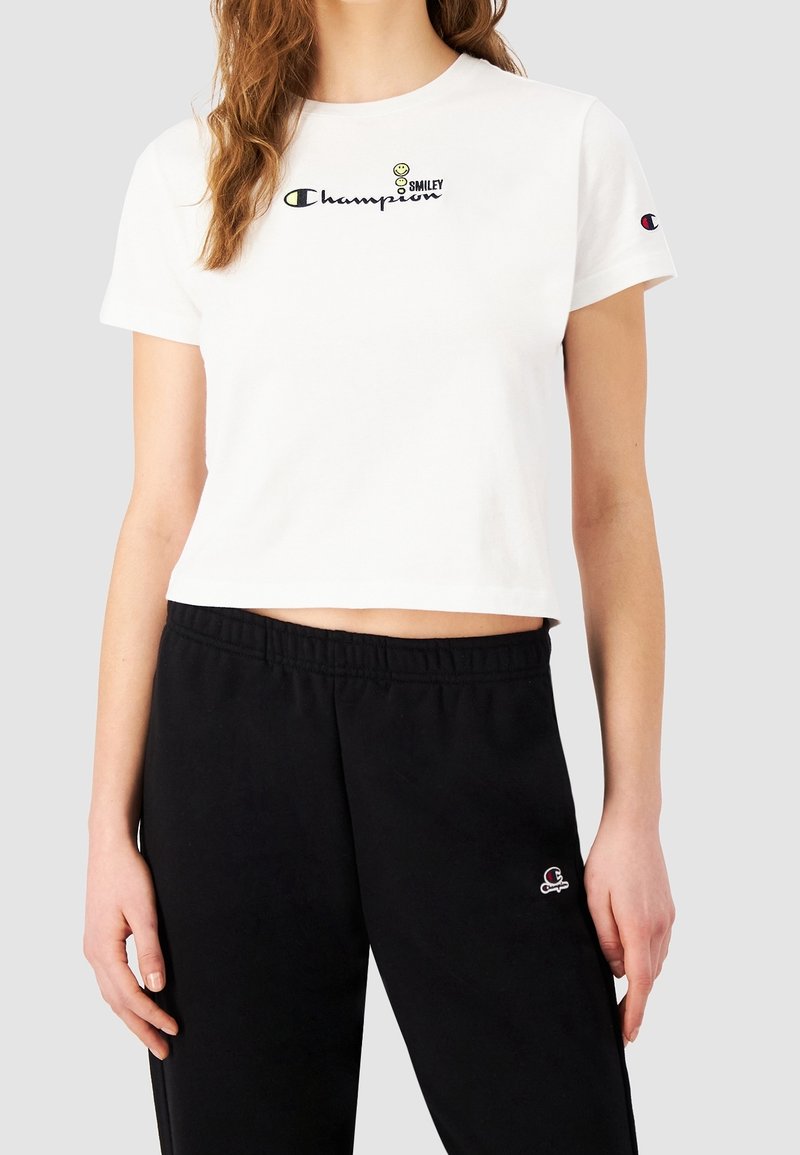 Valkoinen lyhythihainen cropped t-paita, jossa on "Champion"-logo ja Smiley-graafinen kuva; pehmeää puuvillakangasta, pyöreä kaula-aukko, lyhyet hihat. Mustat verkkohousut logoteemalla.