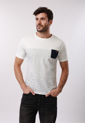 Άνδρας με σκούρα μαλλιά και γενειάδα, που φοράει λευκό T-shirt με σχέδια και μαύρη τσέπη στο στήθος, καθώς και μαύρο τζιν, στέκεται με τα χέρια στις τσέπες.