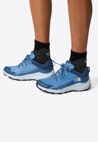 Niebieskie buty sportowe z siateczkową cholewką i gumową podeszwą, zawierające sznurowadła, wzmocniony czubek i logo po boku. W zestawie z ciemnymi skarpetami.