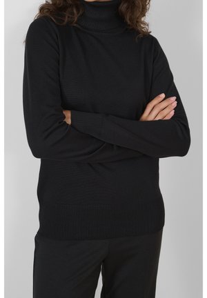 Personne aux cheveux bouclés portant un pull à col roulé noir et un pantalon noir, bras croisés sur la poitrine devant un fond gris.