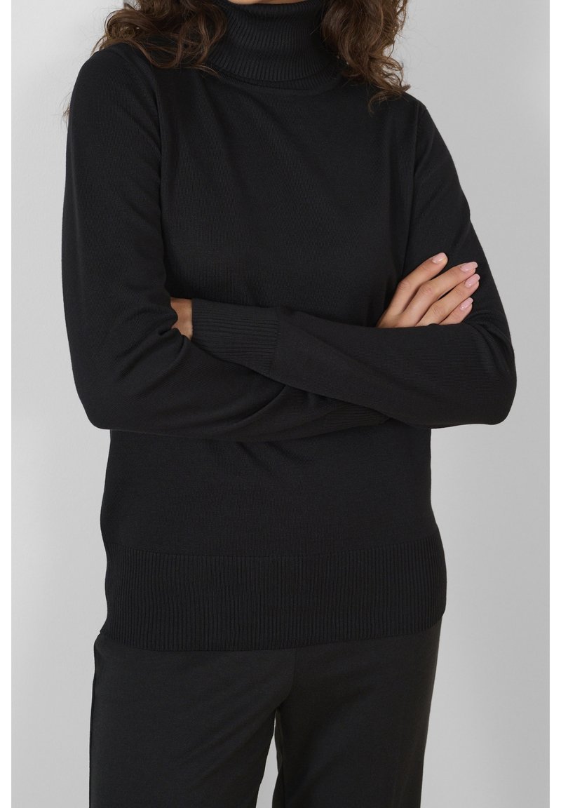 Personne aux cheveux bouclés portant un pull à col roulé noir et un pantalon noir, bras croisés sur la poitrine devant un fond gris.
