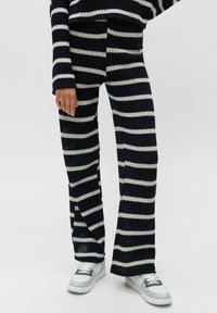 Personne portant un ensemble en tricot noir et blanc à rayures horizontales, avec des baskets blanches et grises, se tenant sur un fond uni.