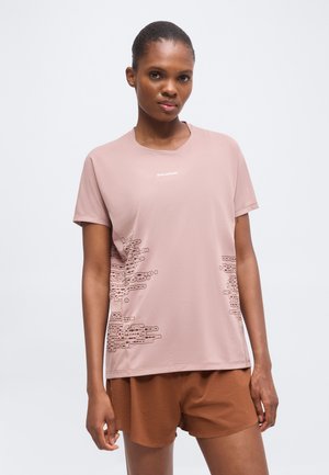 Vrouw draagt een lichtroze T-shirt met korte mouwen met abstracte zijontwerpen en bruine shorts, staand tegen een effen lichte achtergrond.