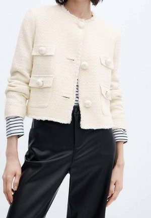 Femme portant une veste courte crème texturée avec de gros boutons sur une chemise rayée et un pantalon noir taille haute.