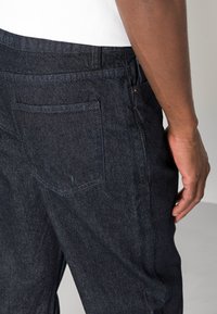 Calças de ganga escuras com um bolso traseiro de design simples, textura de tecido resistente e corte reto, mostradas de uma perspetiva lateral.