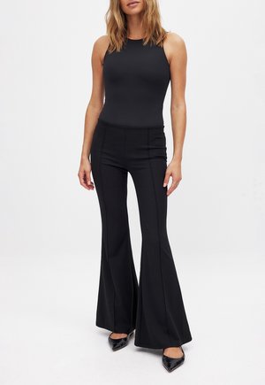 Vrouw met een mouwloos, strak zwart topje, zwarte flarebroek met hoge taille en zwarte pumps met puntige neus, staand tegen een effen achtergrond.