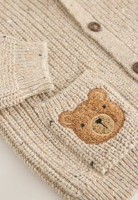 Neule beige kardigaani, jossa on ribbitetty rakenne, esittää ruskean karhun kasvot etutaskussa ja puunapit. Pehmeä, rento muotoilu.