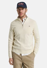 Crème-kleurige zip-up trui met textuurpatroon, geribde manchetten en kraag, over een wit shirt gedragen. Persoon draagt een marineblauwe GANT pet.