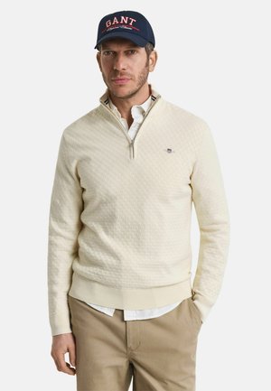 Cremefarbener Reißverschluss-Pullover mit strukturiertem Muster, gerippten Bündchen und Kragen, über einem weißen Hemd getragen. Person trägt eine marineblaue GANT-Mütze.