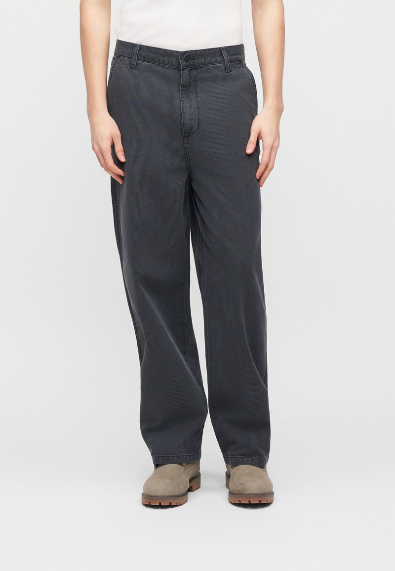 Persona che indossa pantaloni larghi in ciniglia grigio scuro, stivali in pelle scamosciata beige e una maglia bianca, in piedi contro uno sfondo chiaro e uniforme.