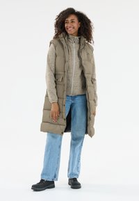 Beige gepolsterte Weste mit hohem Kragen, Fronttaschen und Druckknöpfen, über einem hellgrauen Pullover und blauen weiter geschnittenen Jeans getragen. Schwarze Schuhe.