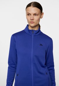 Giacca in pile con zip di colore blu uniforme, presenta un colletto alto, un logo nero sul petto e tasche con zip sui lati.
