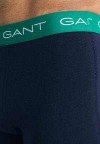 Tumedadest vahtrast kantidark sinise elastse vööga, millel on valge "GANT" logo tekst. Kangas on sile ja veniv.