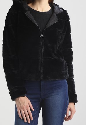 Femme portant une veste noire en fausse fourrure zippée avec capuche et un jean bleu, debout devant un fond clair uni.