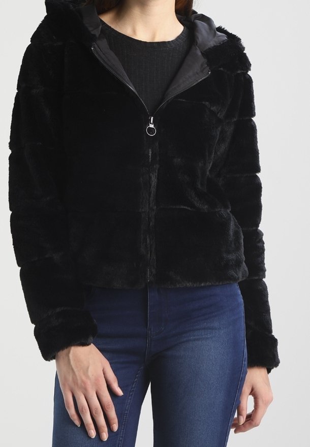 Femme portant une veste noire en fausse fourrure zippée avec capuche et un jean bleu, debout devant un fond clair uni.