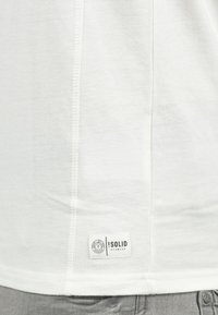 Valkoinen puuvillapaita, jossa on suora leikkaus, litteät saumat ja suorakulmainen logopainatus, jossa lukee "SOLID" alareunassa.