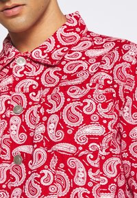 Nahaufnahme einer roten Jacke mit weißen Paisley-Mustern und Metall-Druckknöpfen, getragen von einer Person, die vom Kinn abwärts sichtbar ist.