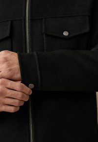 Černá flísová bunda se zipem vpředu, dvěma náprsními kapsami s chlopněmi a detailními cvoky na manžetách.