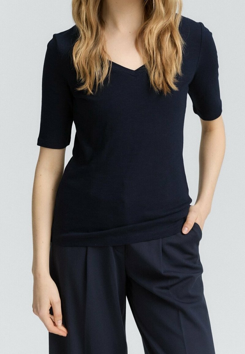 Femme portant un haut noir à manches courtes avec col en V et un pantalon noir, une main dans la poche, avec des cheveux ondulés châtain clair.