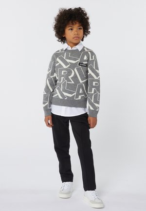 Grijze trui met contrasterende witte grote letters, gelaagd over een witte blouse met kraag. Zwarte broek en witte sneakers maken de outfit compleet.