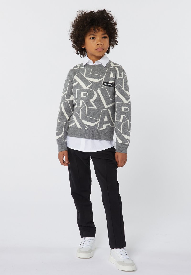 Pull gris avec de grandes lettres blanches contrastantes, superposé sur une chemise blanche à col. Un pantalon noir et des baskets blanches complètent la tenue.