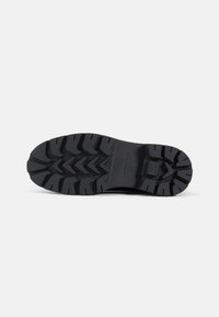 Suela de zapato de goma negra con profundos surcos y una superficie texturizada para mayor agarre, con un diseño minimalista y un acento de la marca cerca del talón.