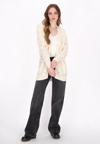 Cremfarbener Strickcardigan mit Rautenmuster, offenes Frontdesign, kombiniert mit hochtaillierten schwarzen weiten Jeans und schwarzen Stiefeln.