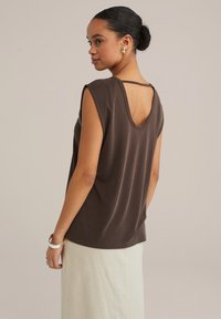 Brun ärmlös topp med låg rygg och rund halsringning, tillverkad av mjukt tyg. Modell som bär den med en ljusfärgad kjol.