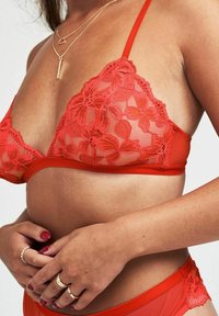 Bralette in pizzo rosso con motivo floreale, coppe in rete trasparente, spalline regolabili e fascia inferiore rossa solida. Le mani della modella poggiano sui fianchi, adornate da anelli.