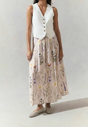 Femme portant un gilet blanc boutonné, une longue jupe fleurie rose clair et des chaussures plates beiges tissées, debout devant un fond uni.