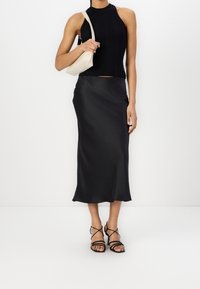 Mujer con una blusa sin mangas negra de canalé, una falda midi de satén negra, sandalias de tacón con tiras negras y llevando un pequeño bolso de hombro beige.