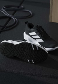 Ζευγάρι ασπρόμαυρα αθλητικά παπούτσια Adidas σε σκοτεινό πάτωμα γυμναστηρίου με ένα μέρος από μία λαστιχένια ταινία αντίστασης και ξύλινο κουτί κοντά.