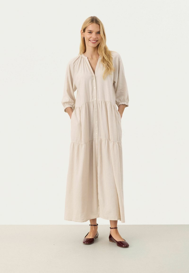 Rochie din in de un bej deschis, cu deschidere pe butoane, talie gathered și mâneci trei sferturi. Prezintă buzunare laterale și un design maxi, fluid.