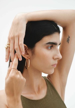 Pendientes de aro de oro y anillos de oro texturizados adornan los dedos. El sujeto lleva una camiseta sin mangas ajustada de color verde oliva, que muestra un diseño simple y un color neutro.
