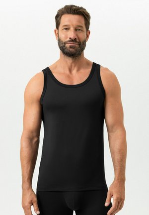 Mann mit Bart, der ein eng anliegendes schwarzes ärmelloses Tanktop trägt und vor einem schlichten hellen Hintergrund steht.