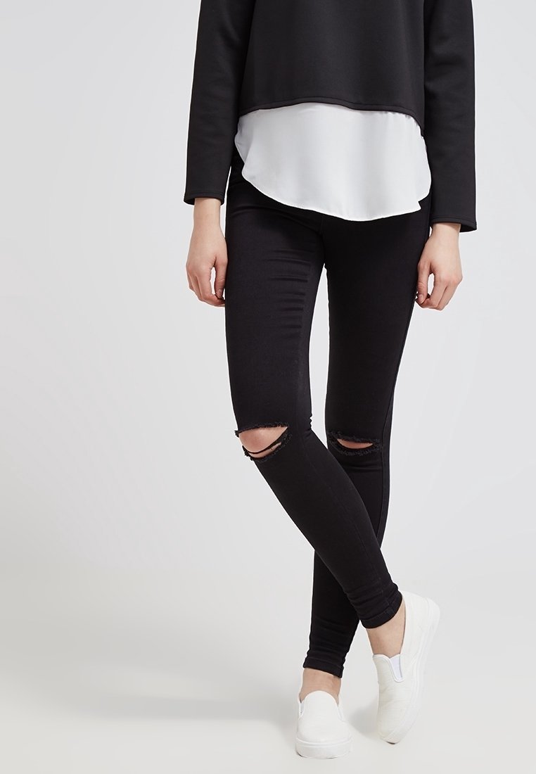Jeans skinny noirs usés avec deux déchirures aux genoux, associés à une blouse blanche en dessous et des chaussures blanches à enfiler. Texture de tissu lisse.
