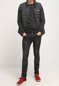 Svart pufferväst över en svart långärmad tröja, kombinerad med mörka jeans och röda sportskor. Vesten har dragkedja och logotyp på bröstet.