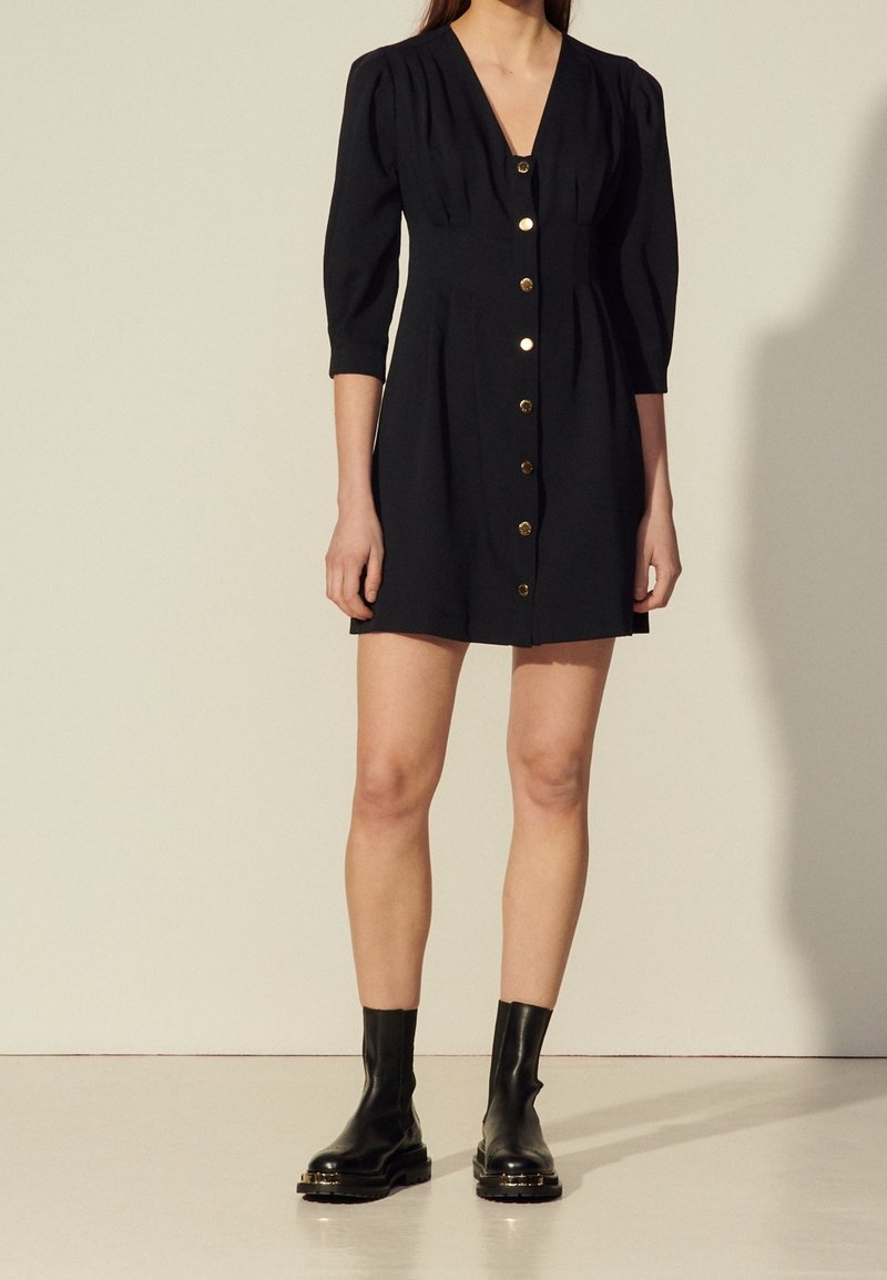 Femme portant une robe noire boutonnée à col en V, manches trois-quarts et bottines noires, debout devant un fond uni.