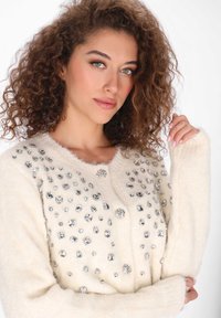 Kremowy kardigan wykonany z miękkiego materiału, z srebrnymi zdobieniami o różnych kształtach i rozmiarach na przodzie. Okrągły dekolt i długie rękawy.