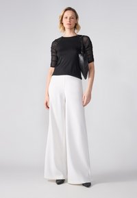 Top negru cu mâneci transparente și drapate; combinat cu pantaloni albi cu talie înaltă și lățime largă; geantă neagră; pantofi negri cu vârful ascuțit.