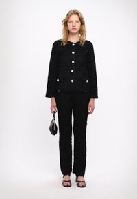 Černý pletený kostým s texturou, který zahrnuje oversized sako s velkými stříbrnými knoflíky a odpovídající kalhoty. Modelka nese malou černou kabelku.