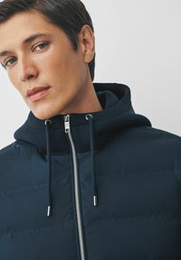 Veste matelassée marine avec capuche, dotée d'une fermeture éclair sur toute la longueur et de détails en métal argenté. Texture lisse avec des coutures matelassées visibles.