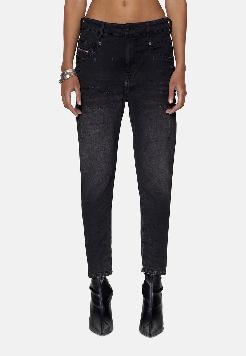 Femme portant un jean skinny noir taille haute avec un léger effet usé, associé à des bottines noires à bout pointu et des bracelets argentés au poignet gauche.