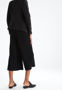 Černé zakázkové sako s saténovými akcenty, spárované s širokými kalhotami z texturované látky. Osoba má na sobě černé nazouvací boty.