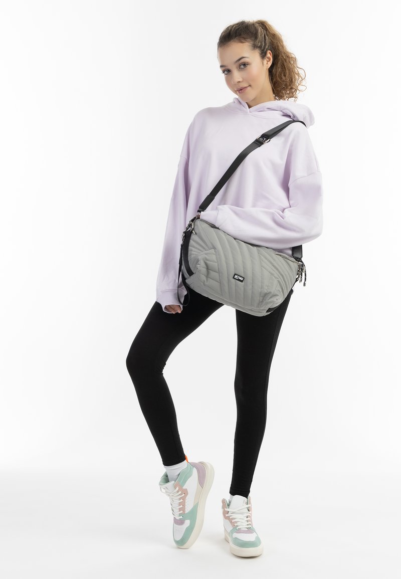 Borsa a spalla grigia trapuntata con chiusure a zip, tracolla regolabile e patch con logo. Il modello indossa un hoodie viola chiaro e leggings neri con sneakers.