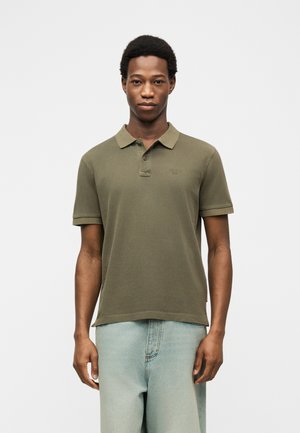 Άνδρας που φοράει απλό χακί κοντομάνικο πόλο και ανοιχτό μπλε τζιν, στέκεται μπροστά σε απλό λευκό φόντο.