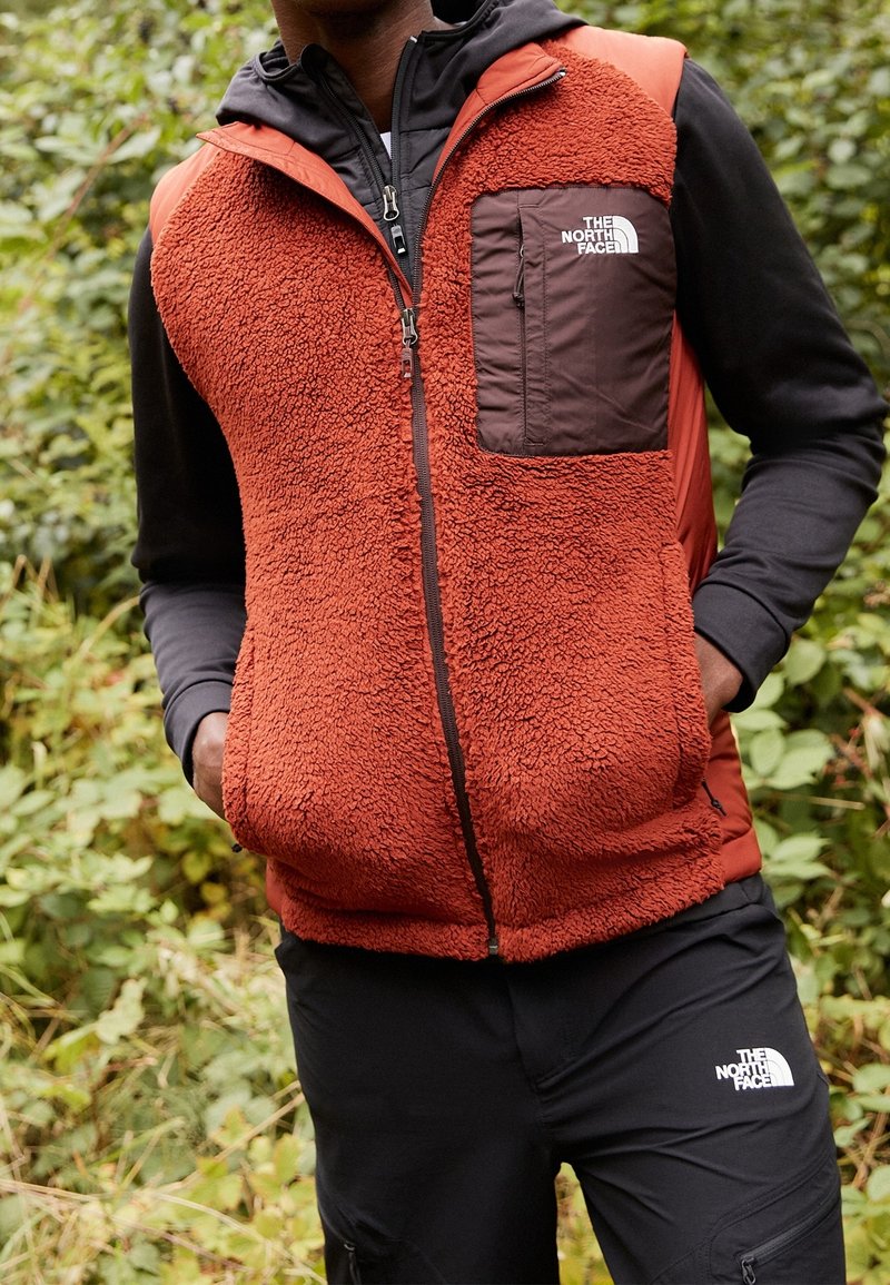 Chaleco de forro polar en color naranja quemado con un bolsillo de nailon marrón, cierre de cremallera y detalles en negro, combinado con pantalones negros que llevan un logo.