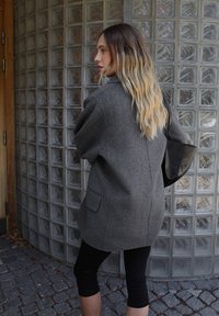 Pilkas vilnos mišinys paltas su laisvu siluetu, su viena kišene, derinamas su juodomis kelnėmis ir laikomas juodu rankiniu. 