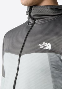 Giacca grigia e azzurro chiaro con colletto alto, tessuto texture, logo sul petto e zip nera. Presenta un design sportivo e una silhouette aderente.