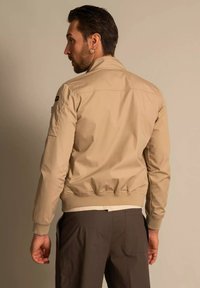 Beżowa bluza bombowca z dopasowanym pasem w talii i mankietami, z boczną kieszenią i gładką fakturą materiału. Minimalistyczny design bez ozdób.